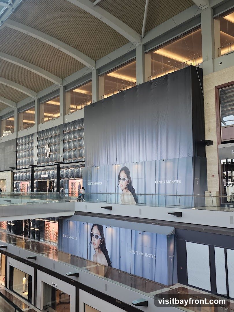 A view of a modern shopping mall interior, showing multiple levels and storefronts. Large advertisements for "GENTLE MONSTER" featuring a model wearing sunglasses are prominently displayed on the upper levels. Below, storefronts for "ZARA" and "MONCLER" are visible. The mall features a contemporary architectural design with high ceilings and glass railings.
