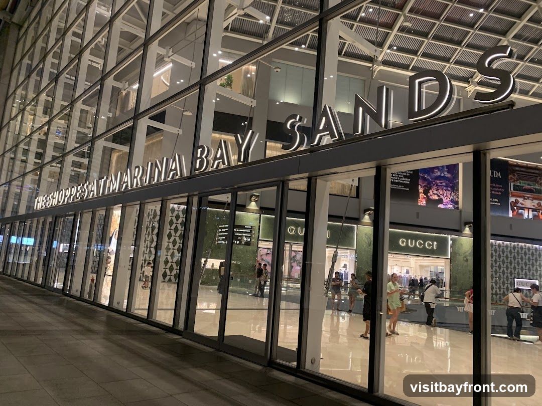 The exterior of a modern shopping mall with large glass windows and doors. The signage "THE SHOPPES AT MARINA BAY SANDS" is prominently displayed above the entrance. Several people are visible inside and outside the mall, and storefronts for luxury brands like Gucci are visible.