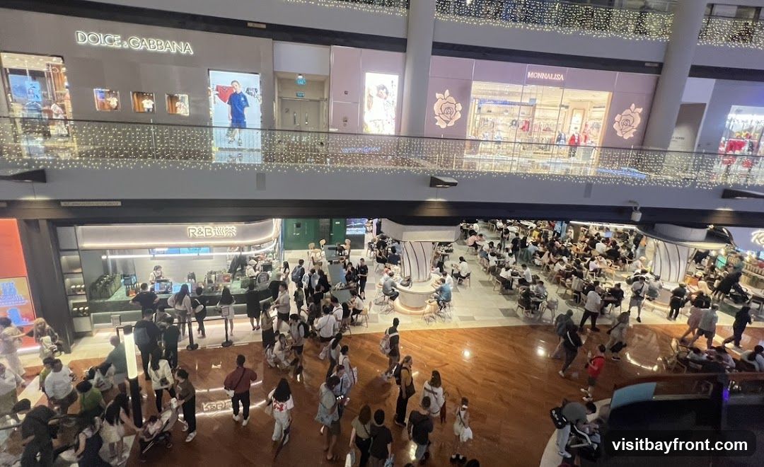 An aerial view of a bustling shopping mall. On the upper level, storefronts for Dolce & Gabbana and Monnalisa are visible, adorned with decorative lights. Below, a busy R&B tea shop is surrounded by a crowd of customers. Further into the mall, a large food court area is filled with people dining at tables. The mall features polished wooden floors and modern architecture.