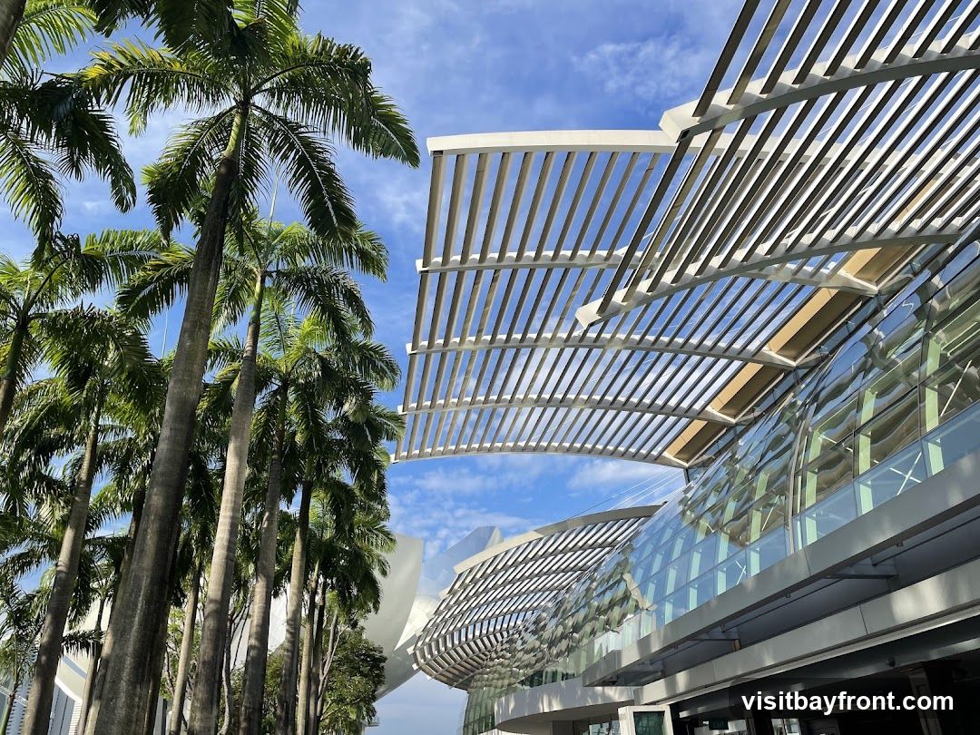 A view of a modern building with a distinctive slatted roof structure and curved glass facade, set against a bright blue sky with scattered clouds. A line of tall palm trees stands to the left, their fronds reaching towards the sky. The architecture appears contemporary and sleek, with a focus on geometric lines and reflective surfaces.