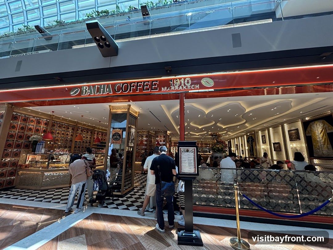 The exterior of a Bacha Coffee shop with its distinctive red and gold branding. The shop front features a large display of coffee tins on the walls, a glass display case with pastries, and seating areas visible inside. The Bacha Coffee logo and "1910 Marrakech" are prominently displayed above the entrance.