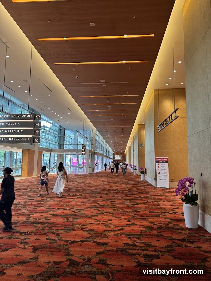 A wide hallway with a patterned carpet and a wooden ceiling. Signs indicate directions to various expo halls and a pick-up/drop-off point. People are walking down the hallway.