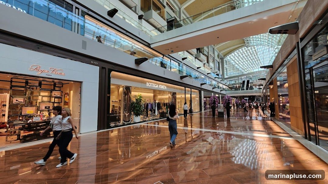 A wide shot of the interior of a modern, upscale shopping mall. The architecture features multiple levels with glass railings and a large, arched glass ceiling that allows natural light to flood the space. The polished, reflective floor mirrors the bright interior. Several luxury brand stores are visible, including Roger Vivier and Bottega Veneta, with their storefronts displaying merchandise. Shoppers are seen walking through the mall, and decorative floral arrangements add to the ambiance.