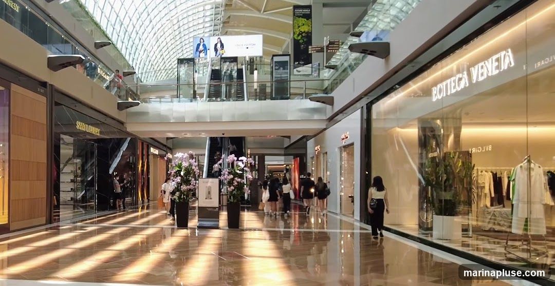 A view of a modern, upscale shopping mall interior with polished floors reflecting sunlight streaming from the ceiling. Several luxury brand stores are visible, including Saint Laurent and Bottega Veneta, with their prominent signage. People are seen walking through the mall and browsing in the stores. The architecture features a high, arched ceiling and multiple levels.