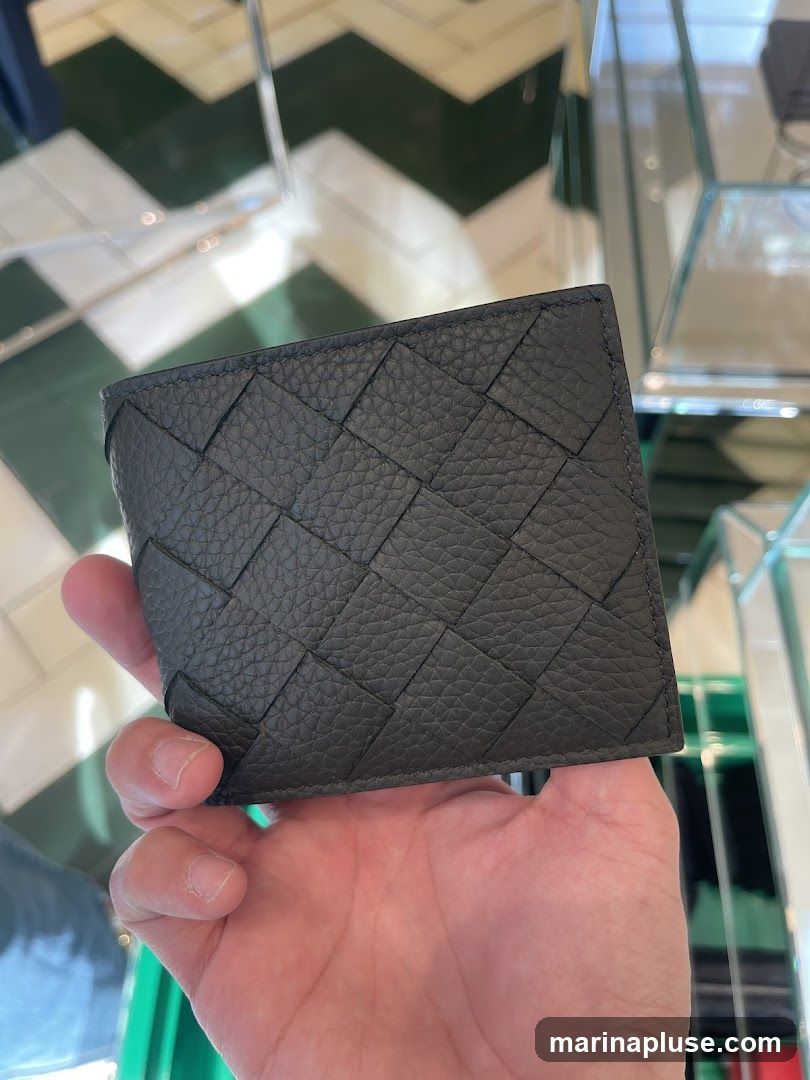 A hand holds a dark gray woven leather wallet. The background shows a checkered floor and glass display shelves.