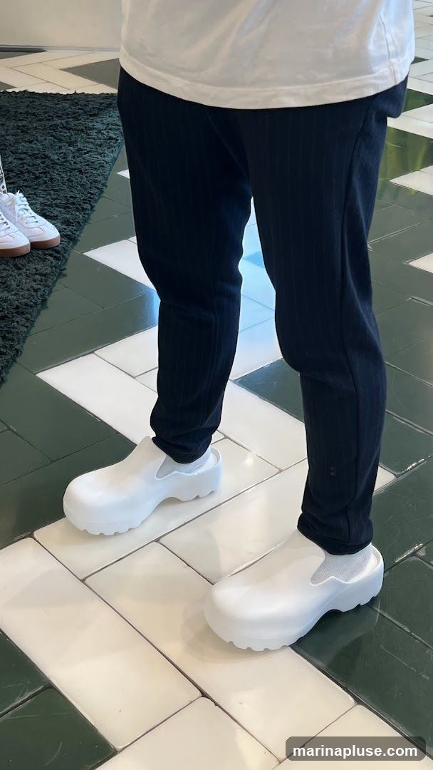 A person wearing white clogs and dark pinstripe pants. Another person wearing white sneakers is visible on the left. The floor has a pattern of white and dark green tiles.