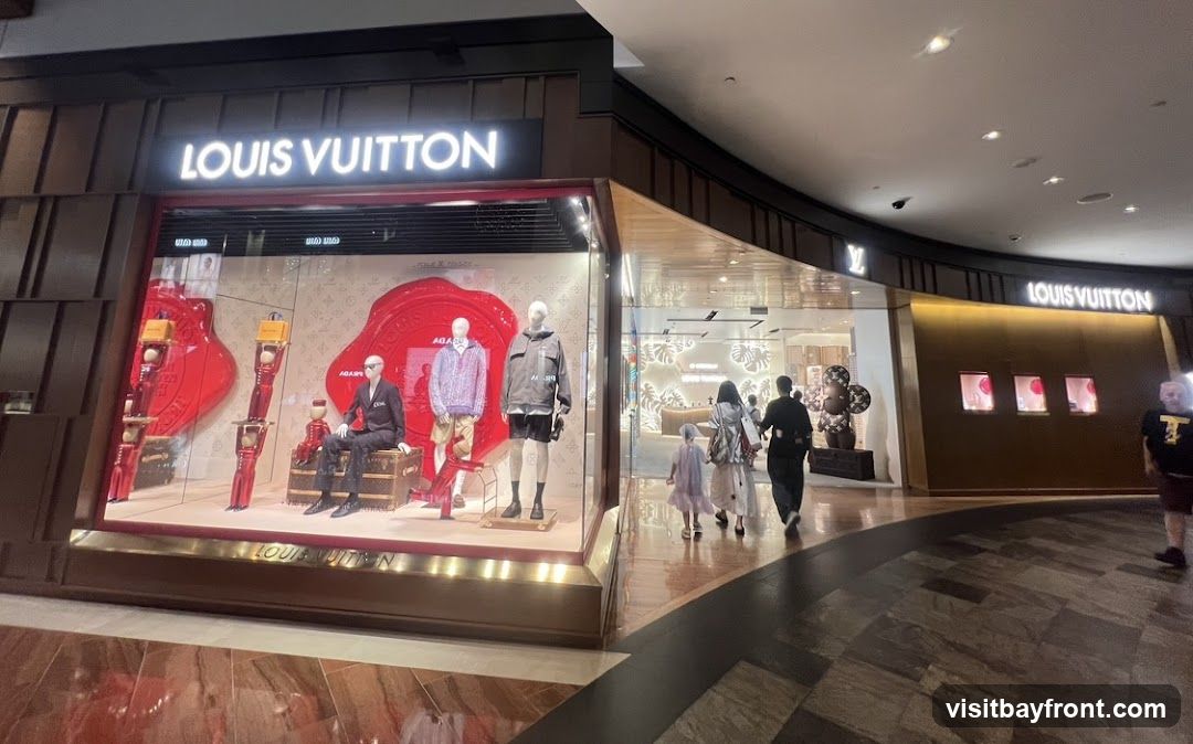 The exterior of a Louis Vuitton store is shown, with a large display window featuring mannequins dressed in fashionable clothing and accessories. The Louis Vuitton logo is prominently displayed above the window and on the facade. People are walking past the store entrance.