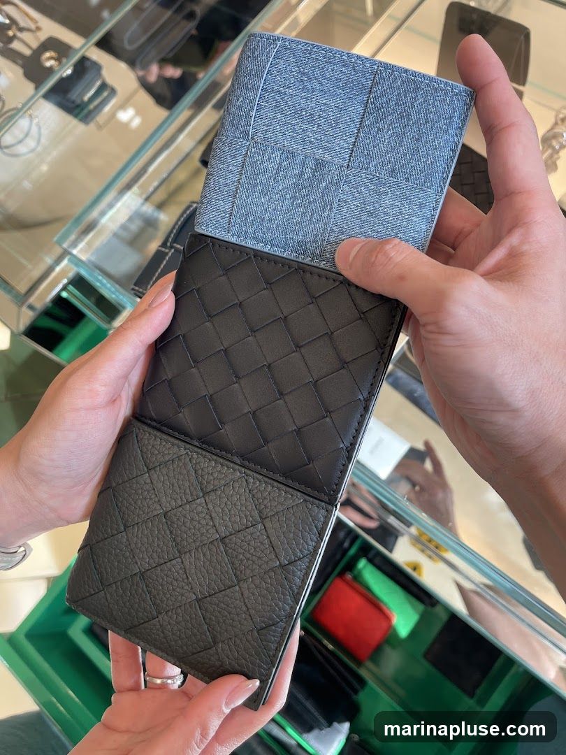 Two hands are holding a wallet with different textures and colors. The top part of the wallet is made of light blue denim-like material, woven in a grid pattern. Below that, a section of black leather is visible, also in a woven pattern. The bottom part of the wallet is a dark gray textured leather, with a diamond-shaped pattern. The wallet is displayed in what appears to be a retail setting, with glass shelves and other items visible in the background.