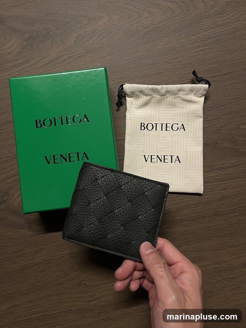 A black woven leather bi-fold wallet is held by a hand, next to its green Bottega Veneta branded box and a drawstring pouch with the Bottega Veneta logo.