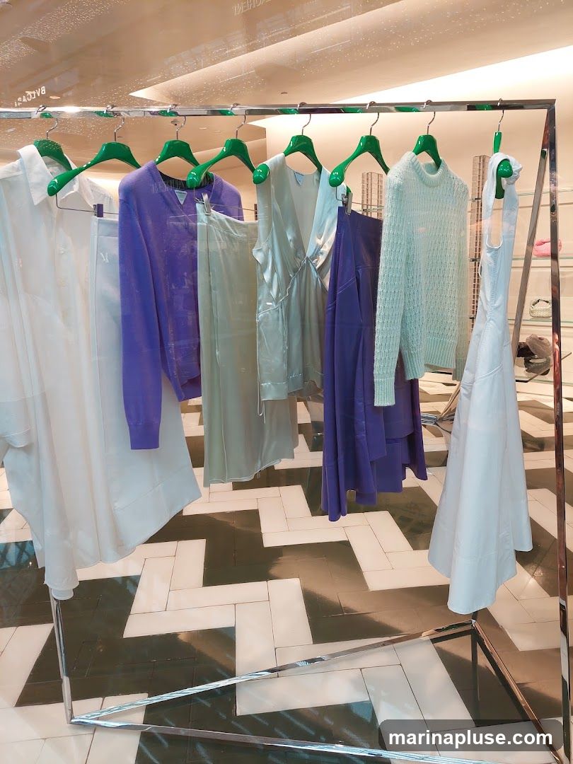 A clothing display featuring several garments on hangers. The garments include a white shirt, a purple sweater, a light green dress, a purple dress, a light blue sweater, and a white dress. The clothes are displayed on a metal rack with green hangers. The background shows a tiled floor with a geometric pattern and a reflection of store lighting.