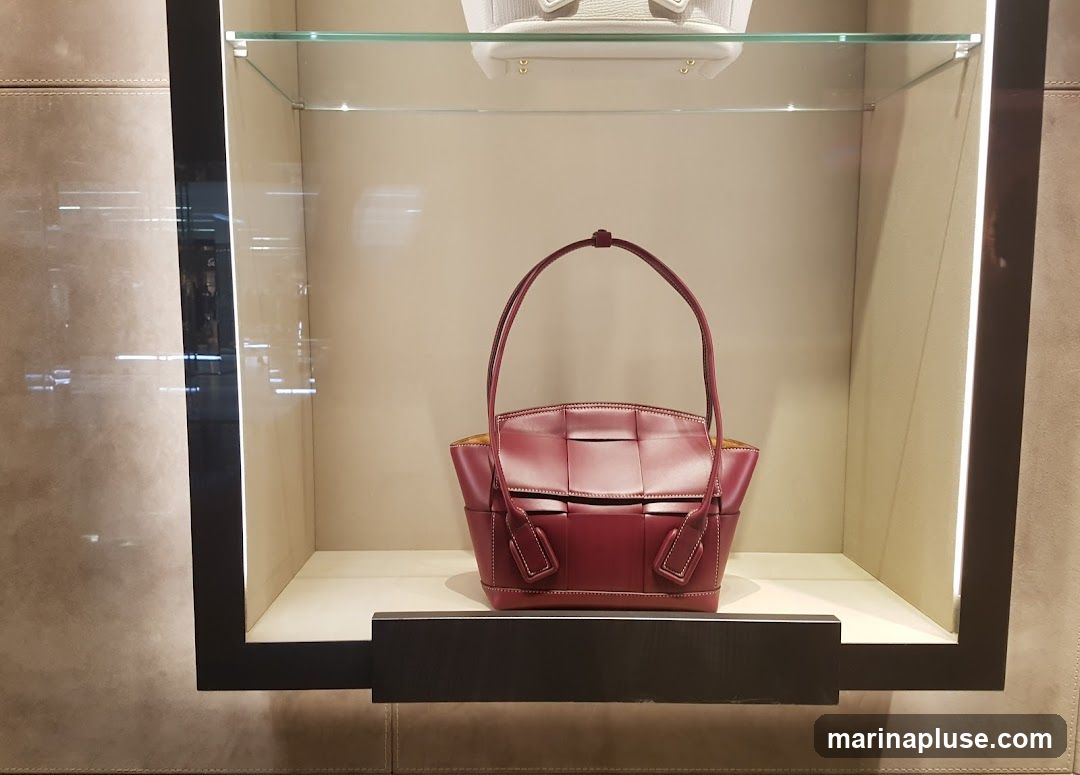 A display case with a burgundy woven leather handbag in the center. Another white handbag is visible on a shelf above. The display case has a dark frame and is lit from within.