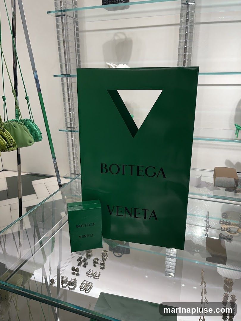 A large green Bottega Veneta shopping bag and a smaller green box with the Bottega Veneta logo are displayed on a glass shelf. Several pairs of earrings and other jewelry items are also visible on the shelf. Green handbags hang from a rack in the background.