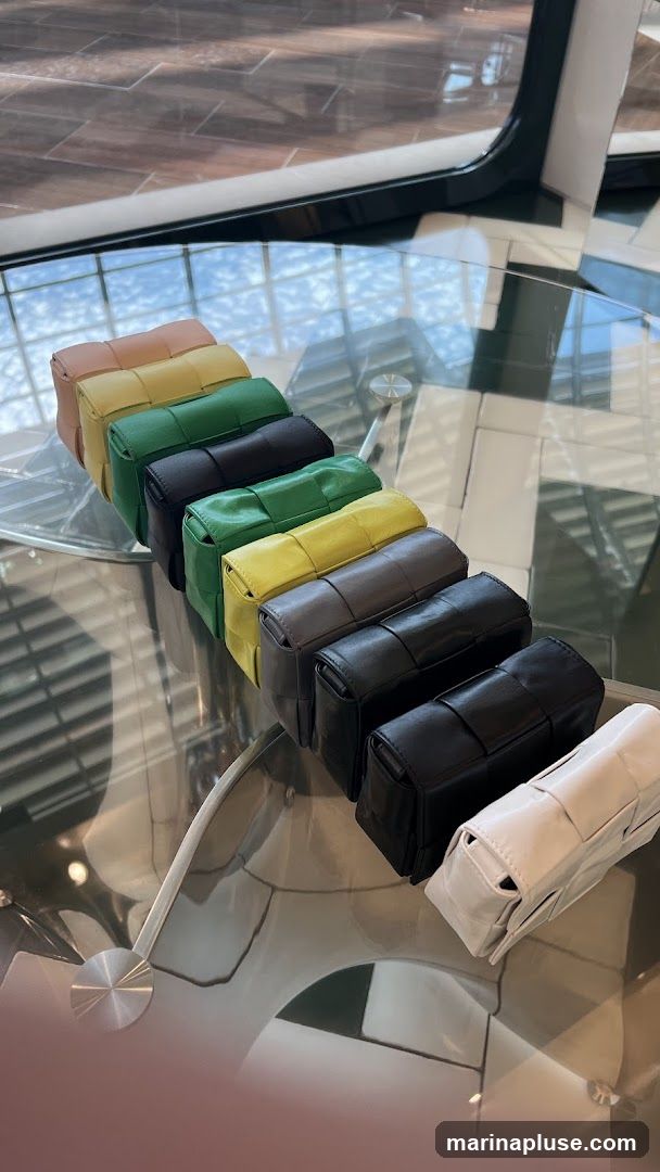 A collection of small, woven leather bags in various colors, including peach, yellow, green, gray, black, and white, are displayed in a row on a glass table. The bags have a distinctive woven texture. The background shows a tiled floor and reflections of the sky and windows on the glass surface.