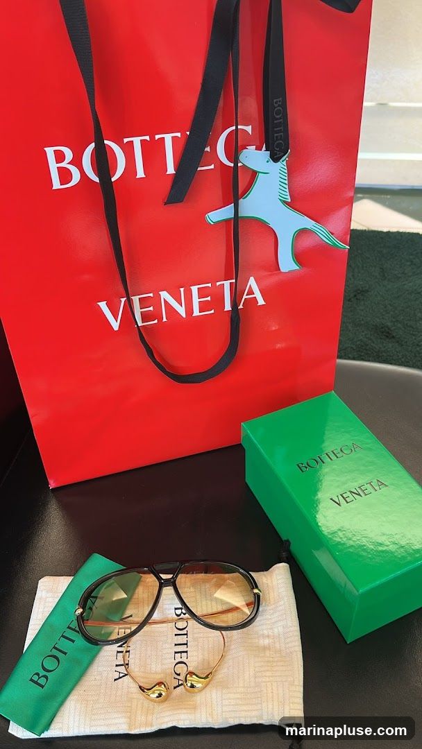 A red Bottega Veneta shopping bag with white lettering is shown. A light blue horse-shaped charm hangs from one of the bag's black handles. In front of the bag are a pair of black aviator sunglasses with gold accents, resting on a beige drawstring pouch. A green Bottega Veneta box is also visible to the right of the sunglasses.