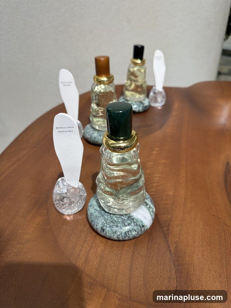 Several small perfume bottles with decorative caps and bases are displayed on a wooden surface. White paper scent strips are placed next to some of the bottles. The labels on the scent strips read "BOTTEGA VENETA COLPO DI SOLE" and "BOTTEGA VENETA".