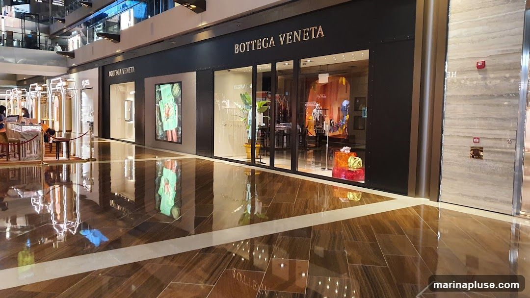 The exterior of a Bottega Veneta store in a modern shopping mall. The storefront features large glass windows displaying merchandise and the brand's name prominently displayed above the entrance. The interior of the store is visible through the windows, showing displays of bags and clothing. The floor in front of the store is highly reflective, mirroring the store's facade and the surrounding mall environment.
