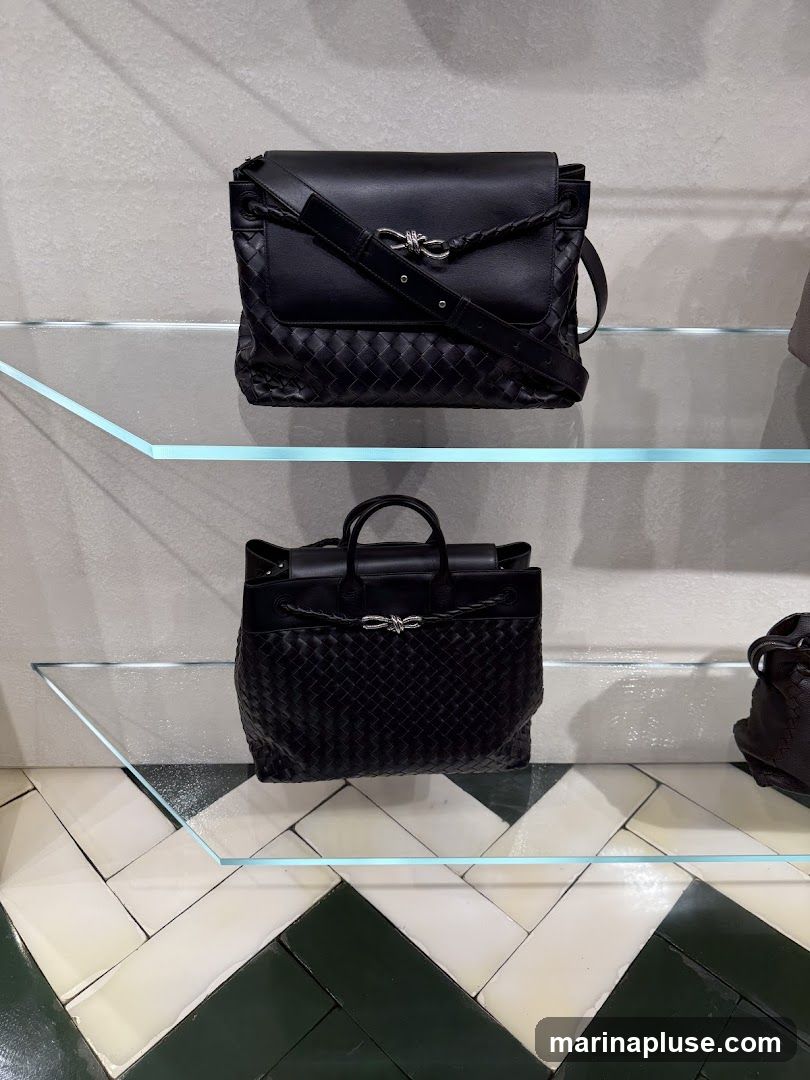 Two black woven leather handbags are displayed on glass shelves. The top bag is a crossbody style with a flap closure and a braided detail. The bottom bag is a tote style with top handles. Both bags feature a distinctive knot-like metal clasp. The background is a textured gray wall, and the floor has a geometric tile pattern in white, cream, and dark green.