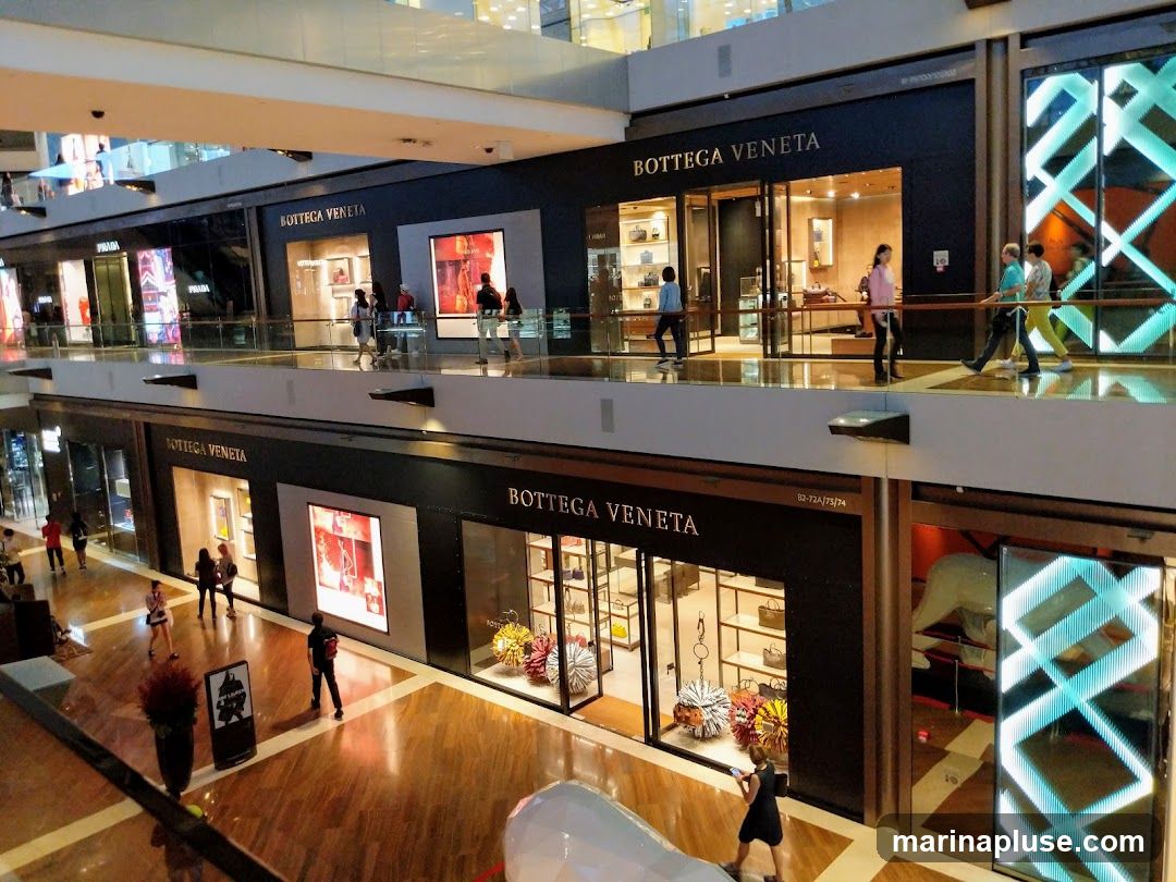 Two levels of a shopping mall with multiple storefronts. The prominent storefronts are for Bottega Veneta, featuring their name in large letters above glass entrances and display windows. The displays showcase handbags and other luxury goods. Other visible storefronts include Prada. People are seen walking on the upper and lower levels of the mall.