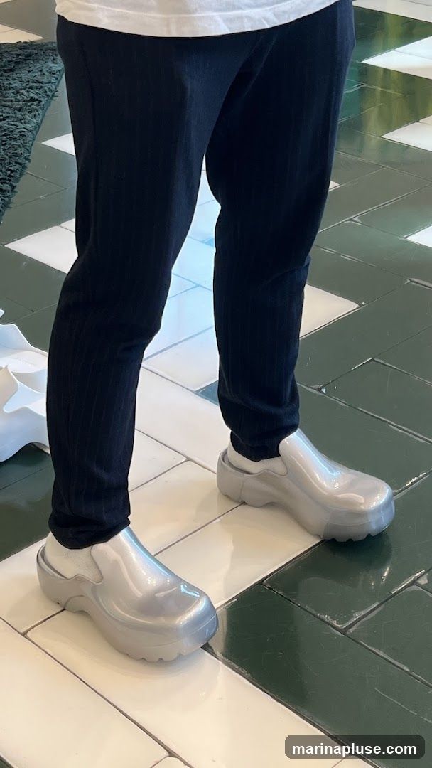 A person wearing black pinstripe pants and white socks is standing on a tiled floor. They are wearing oversized, shiny silver slip-on shoes with thick soles. The floor tiles are a mix of white and dark green, arranged in a checkerboard pattern. A green rug is visible in the background.