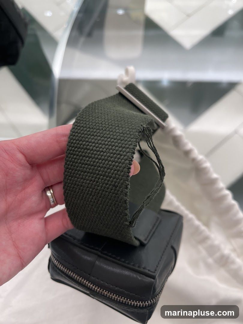 A hand is holding a green canvas strap that is frayed at the end. The strap is attached to a small black leather bag with a zipper closure. The background appears to be a store interior with a checkered floor and reflective surfaces.