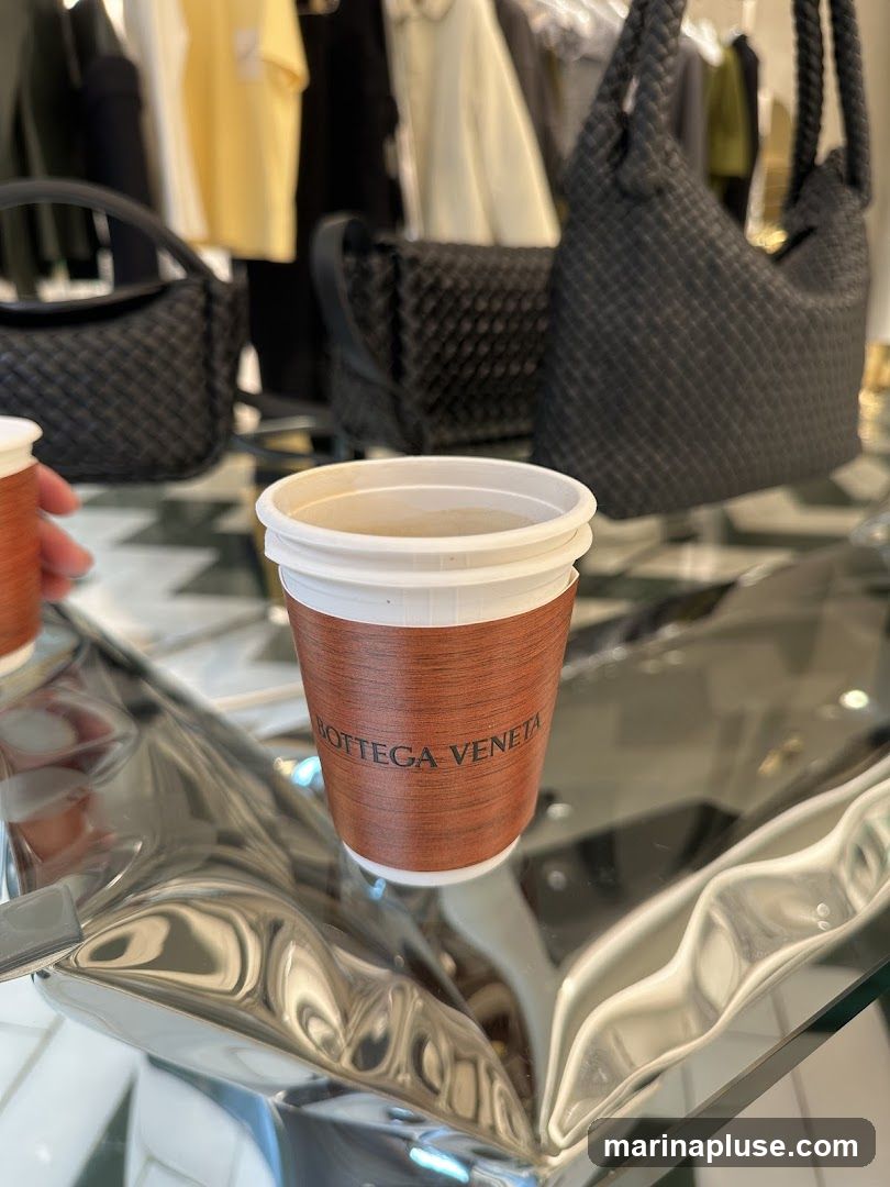 A close-up shot of a disposable coffee cup with a brown sleeve featuring the embossed text "BOTTEGA VENETA". The cup sits on a reflective, wavy surface, likely a table. In the blurred background, racks of clothing and several black woven handbags are visible, suggesting the setting is a retail store.