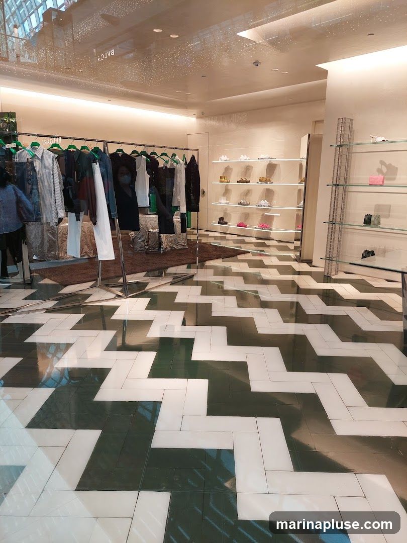 The image shows the interior of a store with clothing displayed on racks and shoes on shelves. The floor has a distinctive zigzag pattern in dark green and white tiles. The lighting is bright and modern, with some reflections visible on the ceiling and glass elements.