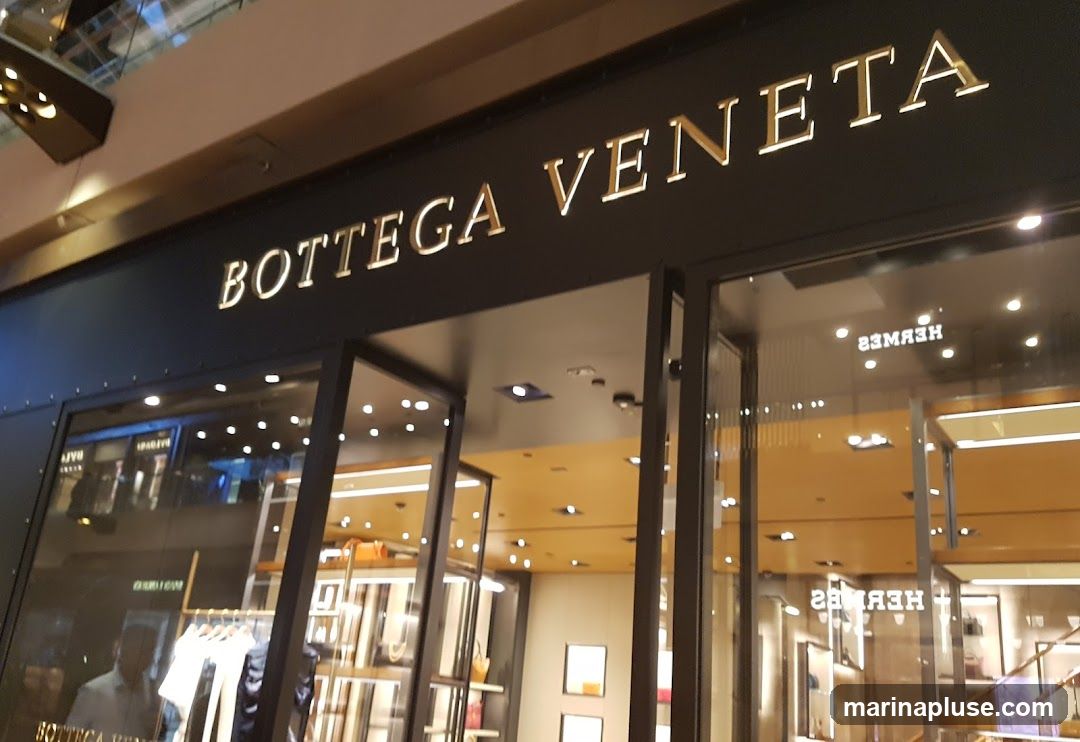 The exterior of a Bottega Veneta store with its name prominently displayed in gold lettering above the entrance. The store features large glass windows and doors, offering a glimpse into the well-lit interior where merchandise is displayed on shelves and racks. Reflections in the glass show other store fronts, including one with the name "HERMES" visible.