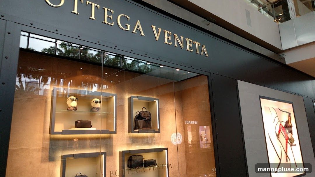 The exterior of a Bottega Veneta store, featuring a large glass window displaying various luxury goods. The store's name is prominently displayed in gold lettering above the window. Inside the display cases, there are mannequins wearing sunglasses, handbags, and backpacks. A framed advertisement with a fashion illustration is visible on the right side of the storefront.