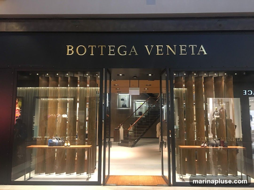 The exterior of a Bottega Veneta store. The store has a dark facade with gold lettering that reads "BOTTEGA VENETA" above a glass entrance. Inside, there are displays of handbags and shoes on wooden shelves. A staircase is visible in the background.