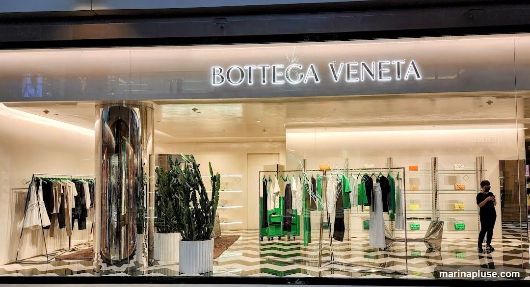 The exterior of a Bottega Veneta store. The store features a large glass facade with the brand name "BOTTEGA VENETA" illuminated in large, white, block letters above the entrance. Inside, clothing is displayed on racks, and handbags are showcased on shelves. A person wearing a mask is visible inside the store. Large potted plants are placed near the entrance. The floor inside has a distinctive black and white chevron pattern.