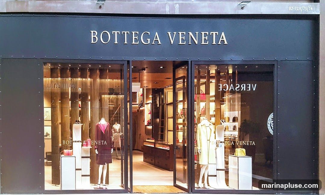 The exterior of a Bottega Veneta store. The name "BOTTEGA VENETA" is displayed in gold lettering above the entrance. The storefront features large glass windows showcasing mannequins dressed in clothing and displays of handbags. The interior of the store is visible, with shelves stocked with merchandise and elegant lighting.