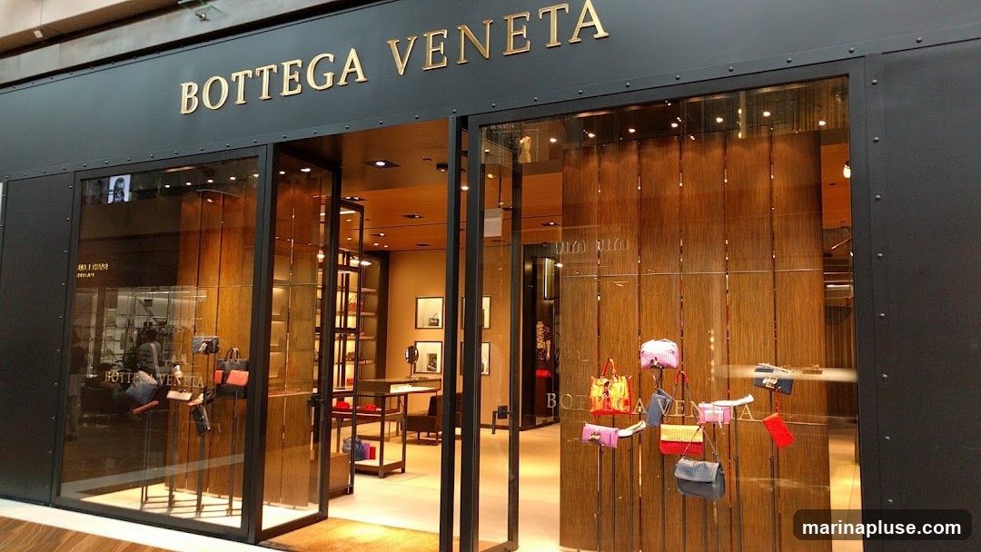 The exterior of a Bottega Veneta store. The store has a dark facade with the gold lettering "BOTTEGA VENETA" above the entrance. The storefront features large glass windows displaying handbags and accessories. The interior is visible through the windows, showing shelves stocked with merchandise and elegant lighting.