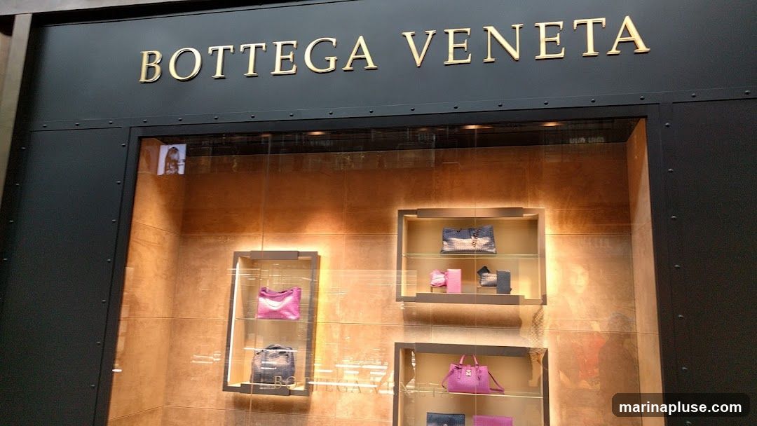 The exterior of a Bottega Veneta store with the brand name displayed in gold lettering above a display window. The window showcases several handbags in various colors and styles, arranged on illuminated shelves. Reflections are visible on the glass.