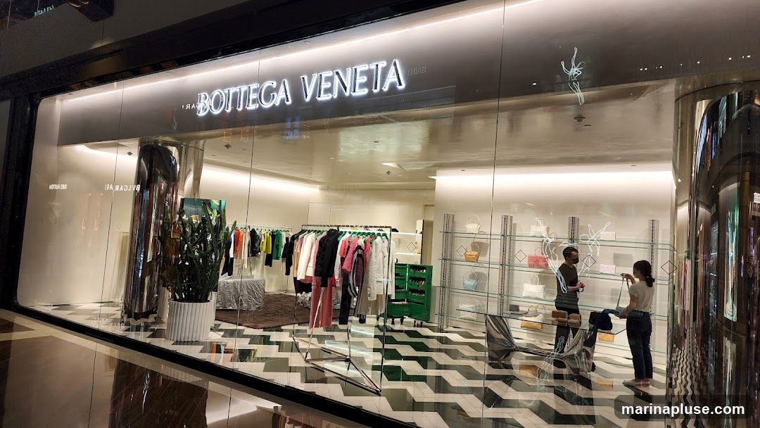 A wide shot of the Bottega Veneta store entrance. The store has a modern and minimalist design with large glass windows and a well-lit interior. Inside, clothing is displayed on racks, and handbags are showcased on shelves. Two customers are browsing in the store. The store's name, "BOTTEGA VENETA," is prominently displayed in illuminated lettering above the entrance.