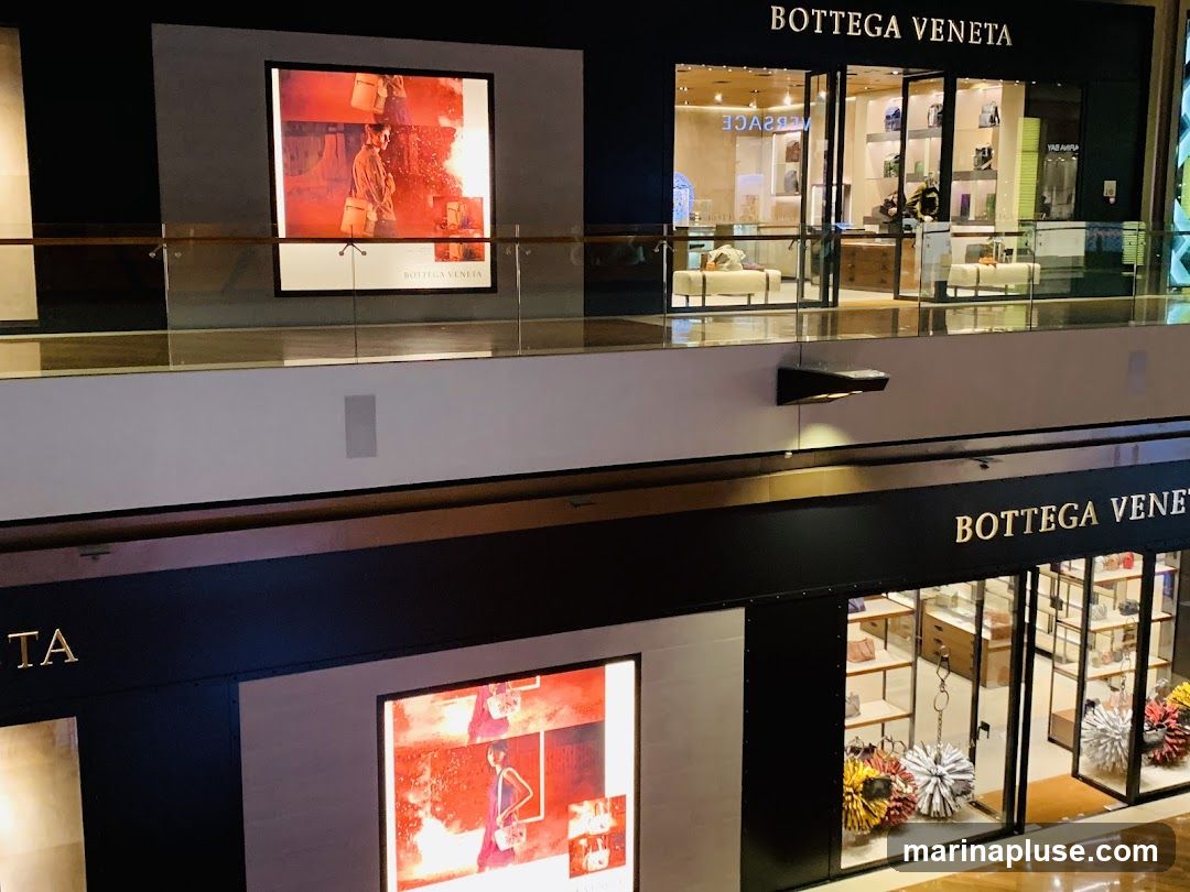 Two storefronts for Bottega Veneta are visible in a shopping mall. The upper storefront has a large display window and a sign above. The lower storefront also features a large display window with merchandise and a prominent sign. Both storefronts are well-lit and showcase luxury goods.