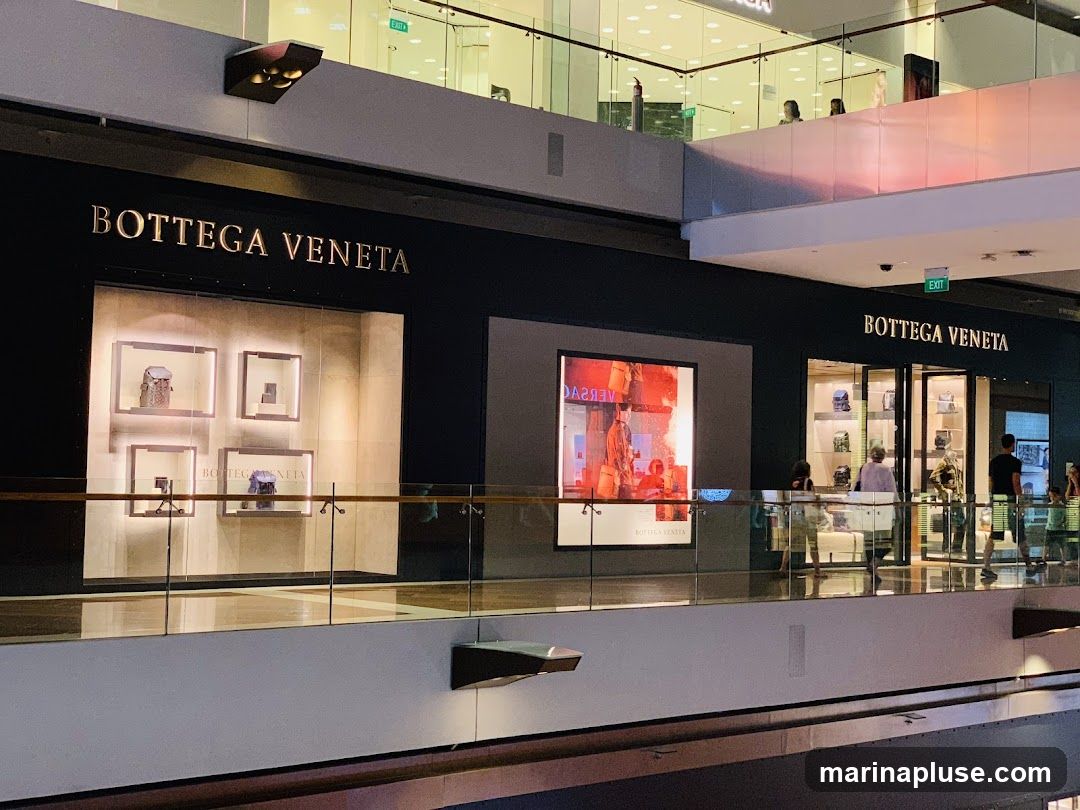 A wide shot of the Bottega Veneta storefront in a modern shopping mall. The store has a dark facade with the brand name prominently displayed in gold lettering. The windows showcase handbags and accessories in illuminated display cases. To the left, a large digital screen displays an advertisement. People are seen browsing inside and outside the store.