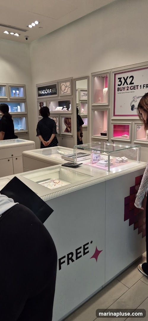 The interior of a Pandora jewelry store with display cases showcasing necklaces, bracelets, and charms. A promotional sign reads "3X2 BUY 2 GET 1 FREE". Several customers are browsing the merchandise.