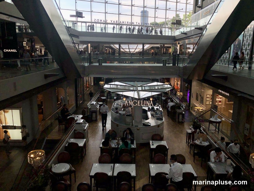 The image shows the interior of a multi-level shopping mall. In the foreground, there is a cafe with tables and chairs. The cafe has a prominent sign that says "ANGELINA". Several people are seated at tables, and staff members are visible. In the background, various luxury brand stores like Chanel and Gucci are visible. The mall has a modern architectural design with glass railings and escalators. People are seen walking on the upper levels.