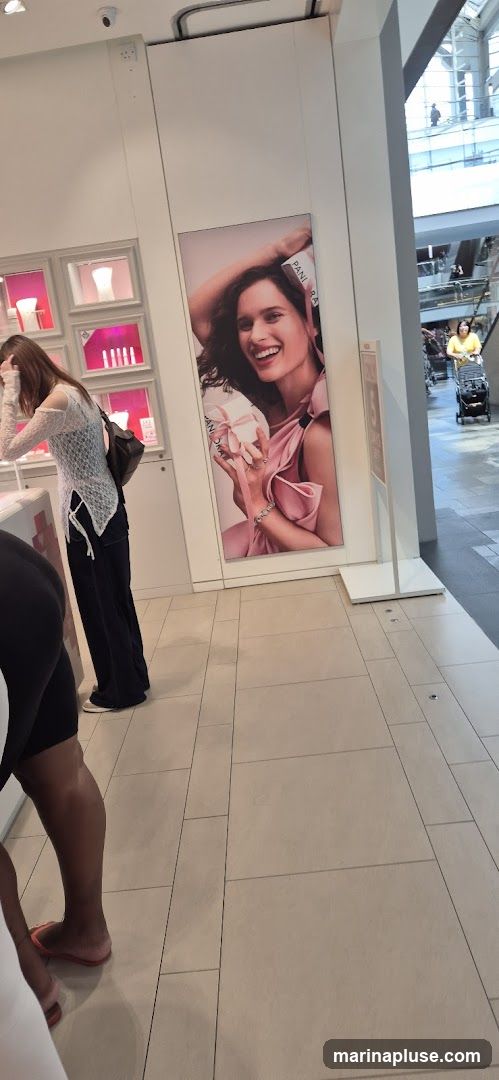 A woman in a white lace top and dark pants looks at a display case in a store. A large advertisement featuring a smiling woman holding a gift box is on the wall behind her. The store appears to be a jewelry store, possibly Pandora, given the branding on the advertisement. The floor is tiled. In the background, other shoppers are visible in a mall-like setting.