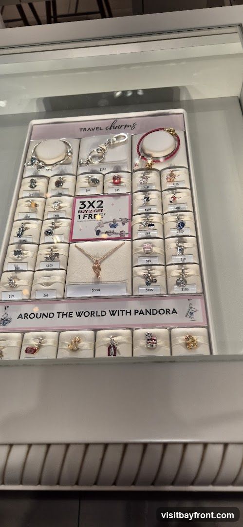 A display case featuring Pandora travel-themed charms and bracelets. The display includes a promotion for "3x2 Buy 2 Get 1 Free". Several charms are priced individually, with prices ranging from $49 to $210. A necklace is also displayed with a price of $334. The display has a banner that reads "AROUND THE WORLD WITH PANDORA".