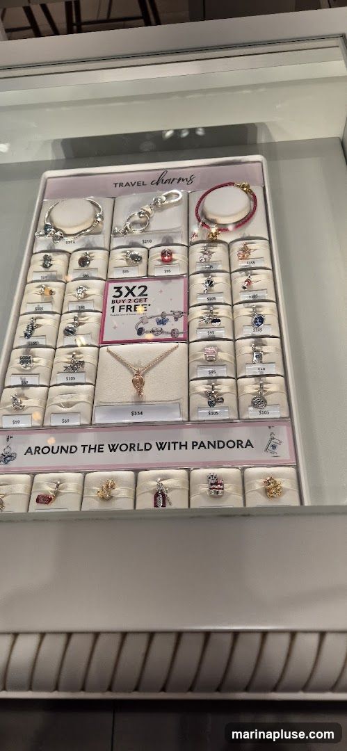 A display case featuring Pandora travel-themed charms and bracelets. The display includes a promotion for "3x2 Buy 2 Get 1 Free". Several charms are priced individually, with prices ranging from $49 to $210. A necklace is also displayed with a price of $334. The display has a banner that reads "AROUND THE WORLD WITH PANDORA".