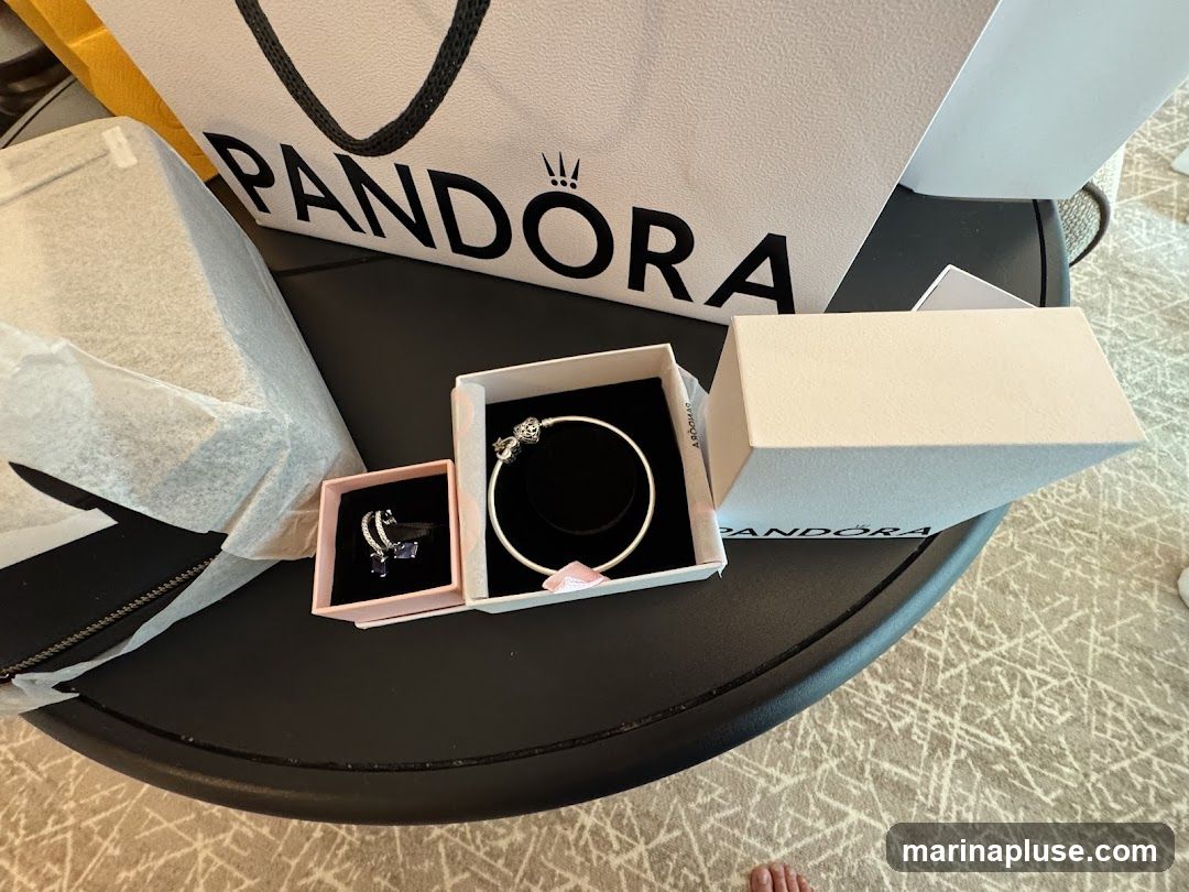 A Pandora shopping bag is visible in the background, with the brand name and logo clearly displayed. In the foreground, several Pandora jewelry boxes are open, revealing a silver bracelet with charms and a ring with a blue stone and diamond accents. Another white Pandora box is closed. The items are arranged on a dark, round surface, possibly a table.