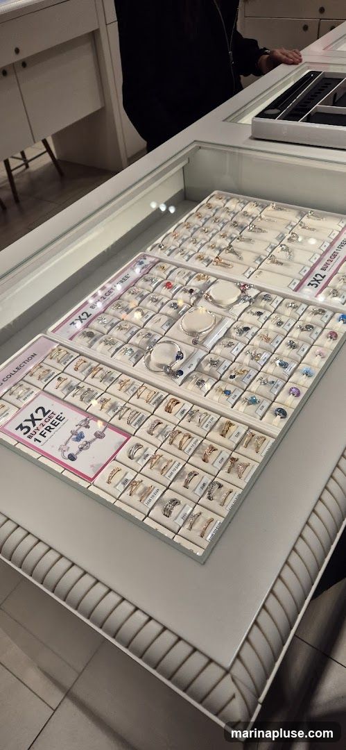 A display case filled with various rings and bracelets. There are promotional signs indicating a "3x2 BUY 2 GET 1 FREE" offer. The jewelry is neatly arranged on white stands within the case.