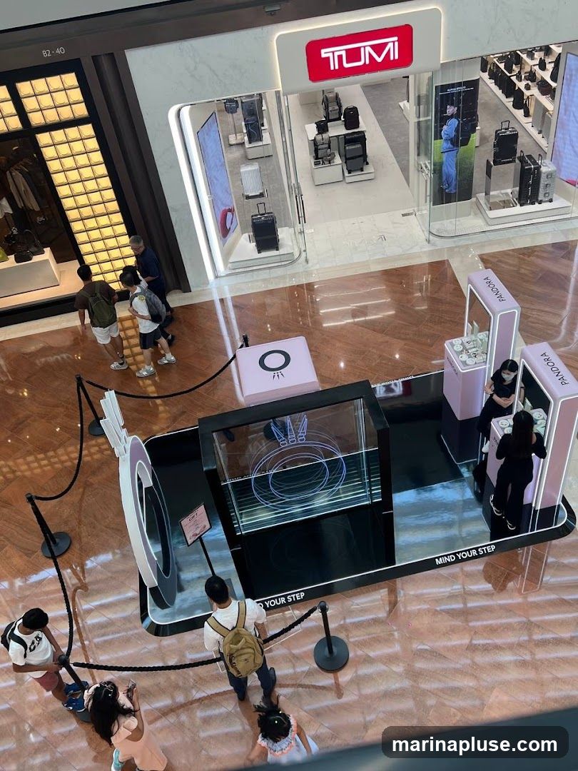 An overhead view of a shopping mall interior. In the foreground, a temporary display with a large ring-like structure and a glass case with a holographic projection is set up, with several people observing it. To the right, a Pandora kiosk is visible with staff attending to customers. In the background, the storefront of a TUMI store is prominent, showcasing luggage and bags. Adjacent to the TUMI store is another shop with a display of clothing and accessories.