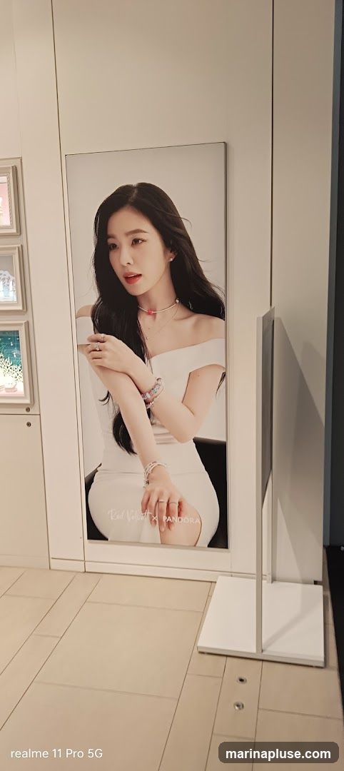 A large advertisement poster featuring a woman in a white dress, wearing jewelry. The text "Red Velvet x Pandora" is visible at the bottom of the poster. The poster is displayed in a store setting, with display cases visible to the left.