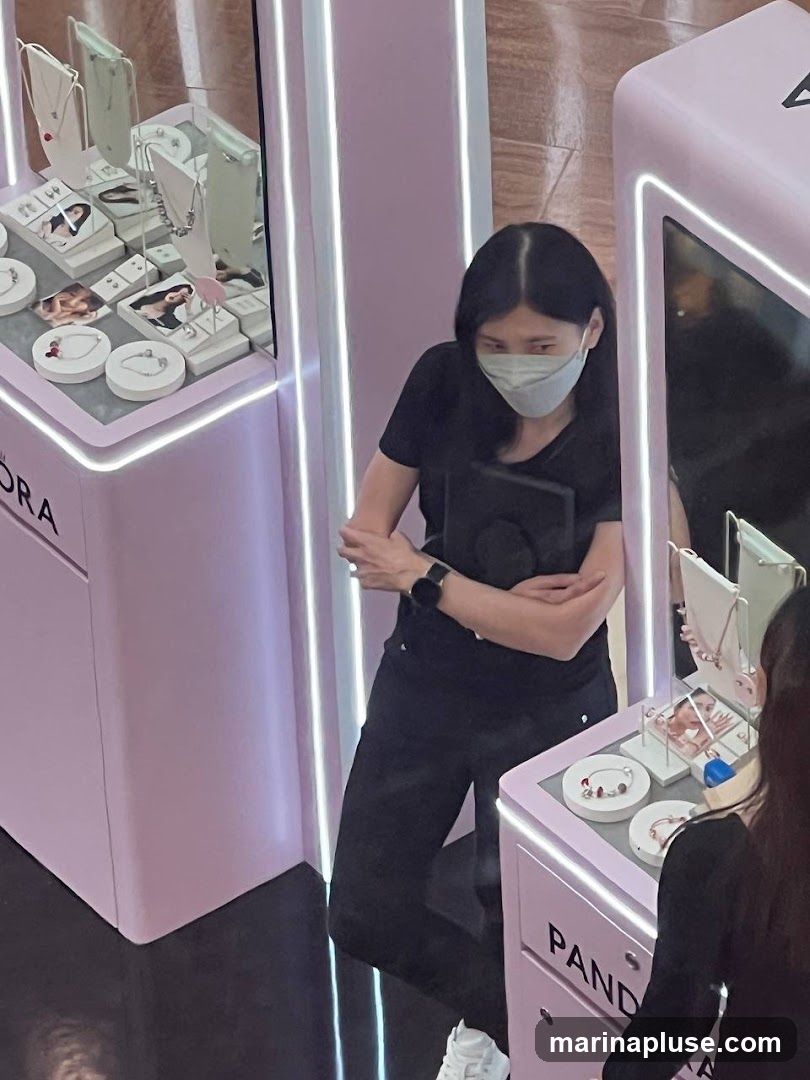 A woman wearing a face mask and a black t-shirt stands in a brightly lit jewelry store. She is looking down and appears to be holding a tablet. The store features pink display cases with jewelry, including necklaces and bracelets. The brand name "PANDORA" is visible on one of the display cases.