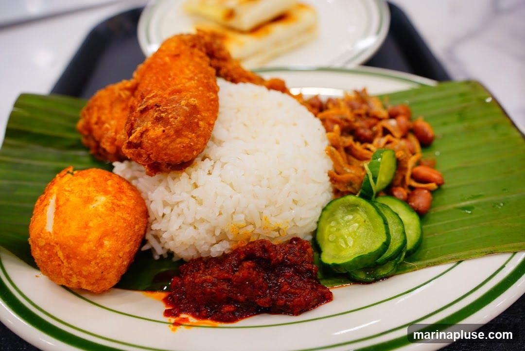 A plate of Nasi Lemak served on a banana leaf, featuring white rice, two pieces of crispy fried chicken, a fried egg, sambal chili paste, anchovies with peanuts, and sliced cucumbers. In the background, a plate with what appears to be roti canai or flatbread is visible.