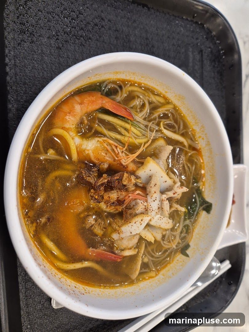 A close-up shot of a bowl of seafood noodle soup. The soup is a rich, orange-brown broth filled with thin noodles. Visible ingredients include large shrimp, sliced fish cakes, and some greens. Fried shallots are sprinkled on top. The bowl is placed on a black tray.