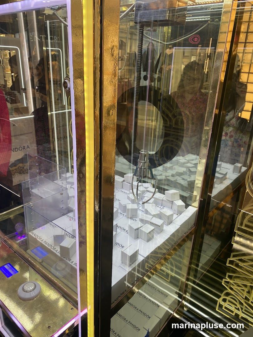 A claw machine filled with small white boxes, many of which are branded with the word "PANDORA". The machine has a gold-colored frame and a clear glass front. The claw mechanism is visible at the top. Reflections of people and store interiors are visible in the glass.