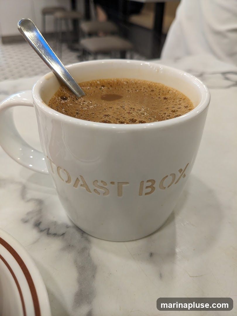 A close-up shot of a white mug filled with coffee, with a spoon resting inside. The mug has the embossed text "TOAST BOX" on its side. The background is blurred, showing what appears to be a cafe setting with tables and chairs.